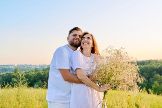 Love and romance of middle-aged couple. Happy hugging man and woman in white dress with bouquet of flowers, summer nature wild meadow. Relationship, holiday, date, wedding, family, people 40s concept