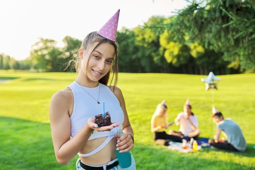 Birthday, teenage girl in festiv hat with cake and candle at outdoor party. Picnic in nature, happy having fun female posing looking at camera. Adolescence, holiday, birthday, celebration, age