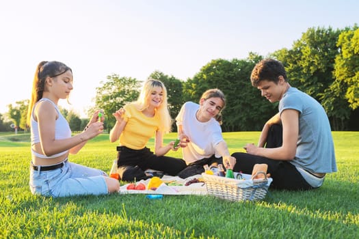 Teens having fun on picnic in park on lawn. Group of teenagers friends resting eating talking on sunny summer day on green grass. Adolescence, friendship, communication, vacations, fun, youth concept