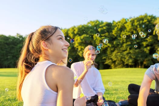 Happy teenage girl 13, 14 years old blowing bubbles. Teenager having fun with friends on lawn in park on sunny day. Fun, summer, vacation, friendship, adolescence concept