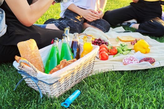 Close-up food and drink for picnic on green grass. Group of young people resting in nature, basket with bread, cheese fruits vegetables snacks juices on tablecloth. Youth, leisure, lifestyle, fun,