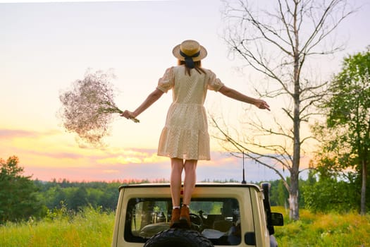 Beautiful woman in straw hat dress with bouquet of flowers back on roof of car, picturesque sunset of wildlife forest meadow. Summer, nature, beauty, people, vacation, weekend, holidays concept