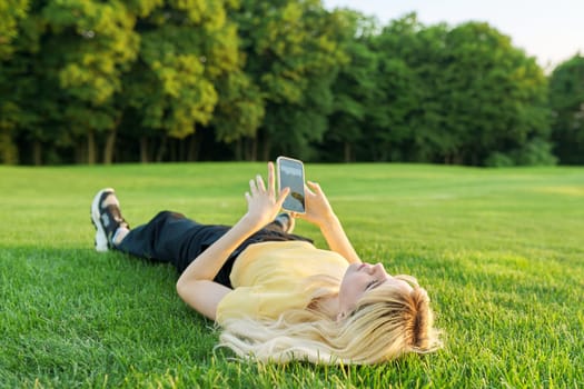 Young teenage woman lying on green grass using smartphone, blonde female resting in park on lawn, in summer sunset light