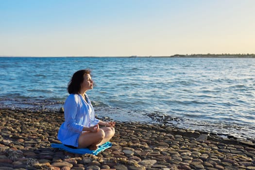 Mature woman sitting in lotus position meditating on the beach. The female enjoys sea nature, the setting sun, summer vacations. Lifestyle, tourism, travel, beauty, health, mature people concept