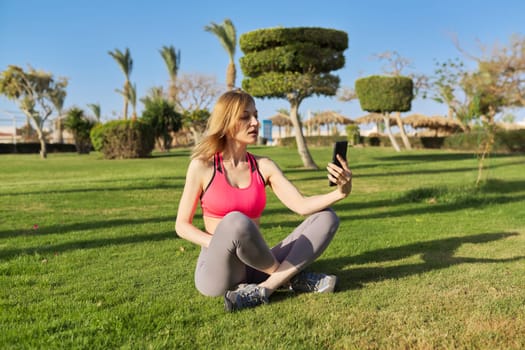 Middle aged active woman doing morning workout, sitting in park on grass, using smart phone for video exercising, chatting, video call.