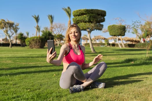Middle aged active woman doing morning workout, sitting in park on grass, using smart phone for video exercising, chatting, video call.