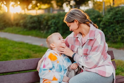 mother and son sit on a park bench in the rays of the setting sun. the concept of a family. Mother's Day. beautiful girl (mother) with a boy (son) in the park in the park are sitting on a bench at sunset.