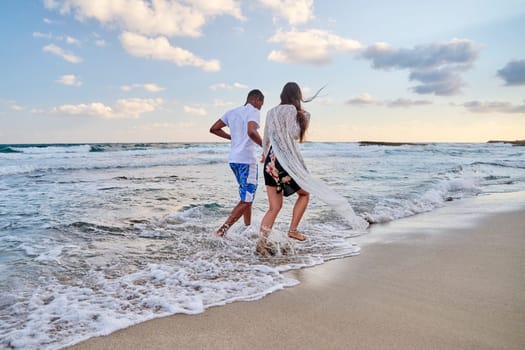 Happy couple running together on the water, on the beach on a summer day, back view. Active young man and woman enjoying sea, nature, vacation, love, relationship, communication concept