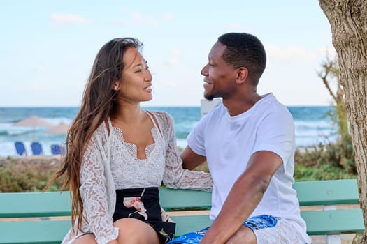 Young multicultural couple smiling talking sitting on bench, sea background. Beautiful asian woman and african american man together outdoor, sea vacation together, love, relationship, communication