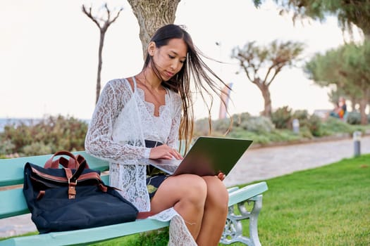 Beautiful young Asian woman using laptop sitting on an outdoor bench. Focused serious female looking in laptop screen, exotic nature sea background. Freelance, remote work study, technology, lifestyle