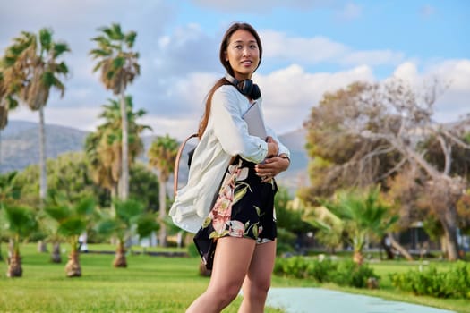 Portrait of young woman with backpack, headphones and laptop in her hands, summer park with palm trees background. Beautiful Asian female student, freelancer, blogger, learning, working remotely