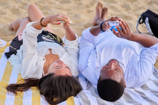 Young multicultural couple lying on beach using smartphones. Relaxing tourists lying on beach towels looking at smartphones. Lifestyle, technology, tourism, travel, nature, leisure, people concept