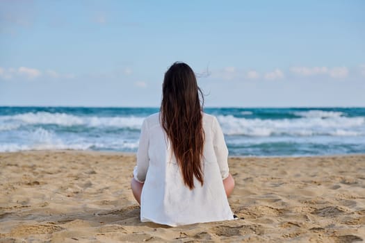 Beautiful brunette in white with long hair sitting in lotus position on sea sandy beach with back looking out horizon. Beauty, tranquility, relaxation, nature, vacation tourism, health. Sky copy space