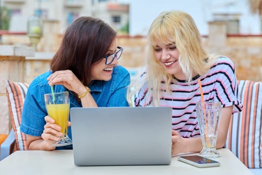 Smiling mother and teenage daughter looking in laptop screen together. Mom and teen girl resting together in outdoor cafe. Parent and child teenager, family, relationship, lifestyle, leisure concept
