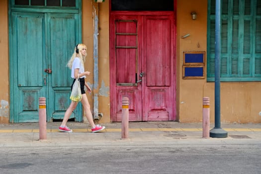 Young teenage woman walking along street of old city, colored wooden vintage doors background. Fashionable blonde in headphones with mesh bag of bananas, profile view. Fashion, style, trends, tourism, travel, lifestyle, youth