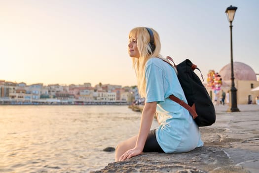 Young blonde woman with long hair in headphones with backpack sitting on city sea promenade at sunset, listening to music, enjoying freedom, happiness, joy, holiday, relaxation, nature, beauty, copy space