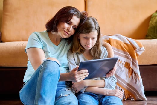 Middle-aged mom and preteen daughter relaxing at home together, looking into a digital tablet. Parent child relationship, family friendship, weekend, lifestyle, home leisure, entertainment concept