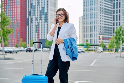 Middle aged confident business woman with suitcase, urban background. Smiling mature woman with smartphone luggage, looking at camera. Business trip, travel, city, work, lifestyle, people 40s concept