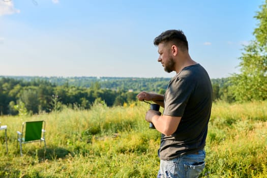 Middle aged bearded man grinding and brewing coffee while relaxing on a sunny summer day, copy space. Camping, summertime, wild meadow, nature, 40s people concept