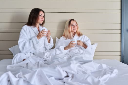 Happy mature mother and teenage daughter drinking coffee, talking, smiling. Women in white bathrobes, resting in bed, vacation together. Communication between parent and adolescent child, family