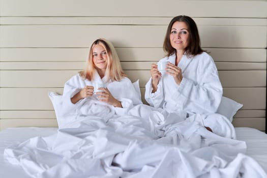 Happy mature mother and teenage daughter drinking coffee, talking, smiling. Women in white bathrobes, resting in bed, vacation together. Communication between parent and adolescent child, family