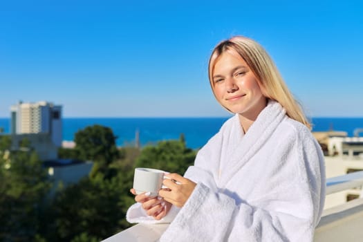 Summer vacation at seaside resort. Happy young woman in white bathrobe enjoying cup of coffee and sunny landscape of sea resort on balcony terrace of hotel, blue sky copy space
