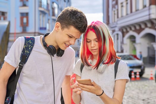 Teenagers students guy and girl with backpacks and headphones look in smartphone screen, talk, city street background. High school and college, communication, youth concept