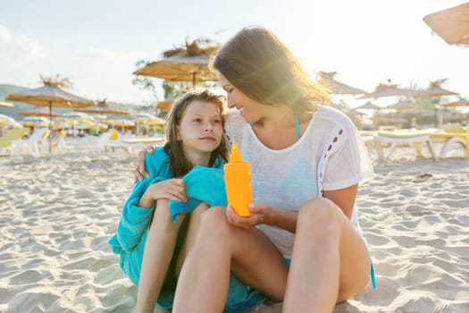 Mom applying sunscreen lotion to daughter's skin, sandy beach summer straw umbrellas background. Family, vacation together, care, love parent and child