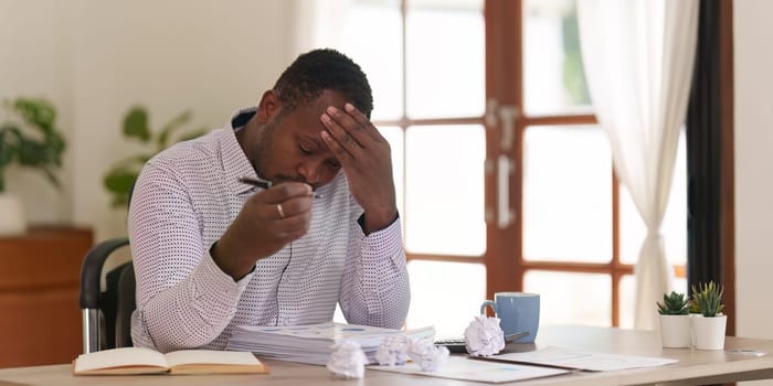 Tired American African business man in stress works with many paperwork document. migraine attack. Freelance, work from home.