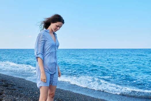 Young beautiful woman in a blue shirt with wet hair on the beach looking down, sea sky nature background for copy space