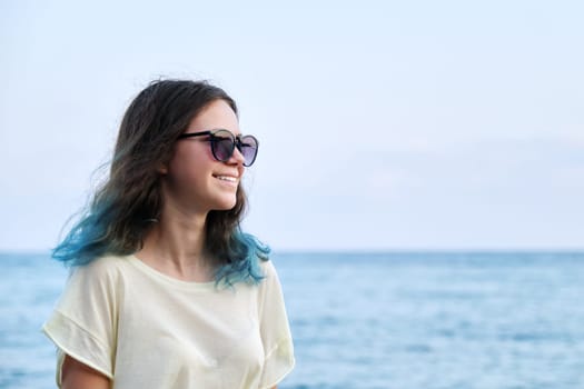 Beautiful teenager girl in sunglasses with dyed blue hair on sea beach, nature sea sky background for copy space. Adolescence, beauty, vacation, weekend concept