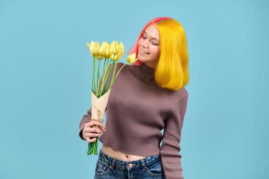 Beautiful smiling trendy teen model with bouquet of yellow tulips on blue studio background. Girl with colored dyed hair, in jeans sweater, enjoying flowers