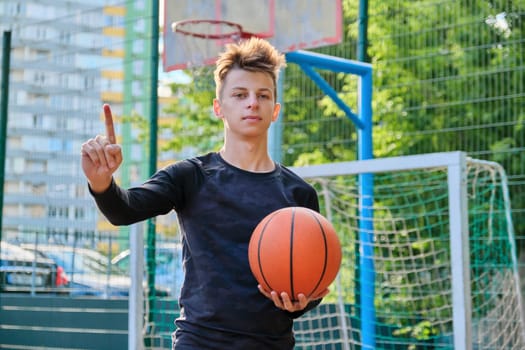 Serious kid teenager with basketball ball showing thumb up attention sign hand gesture, street basketball court background