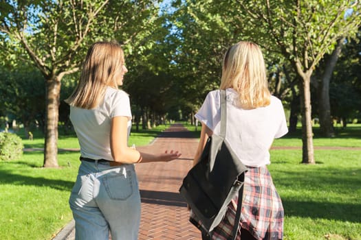 Two happy smiling talking girls teenagers students walking together, back view, young women with backpack, sunny day in the park background