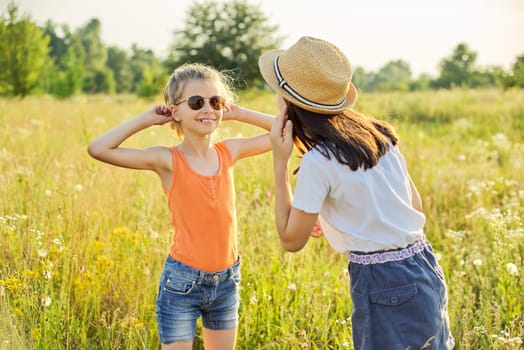 Children are having fun in nature. Two girls laugh, look at themselves in the mirror of sunglasses, sunny summer meadow with grasses background. Childhood, fun, happiness, summer concept