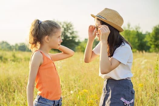 Children are having fun in nature. Two girls laugh, look at themselves in the mirror of sunglasses, sunny summer meadow with grasses background. Childhood, fun, happiness, summer concept