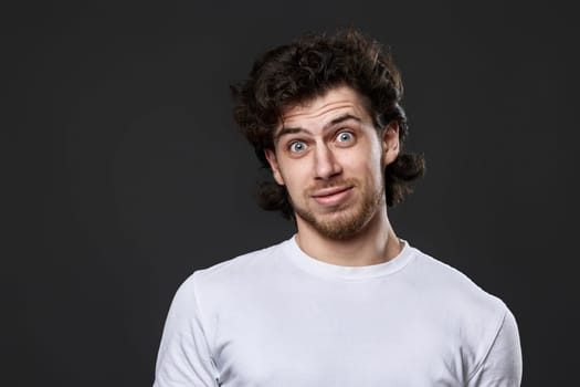 puzzled young bearded man in casual white t-shirt looking at the camera on dark gray background. great idea