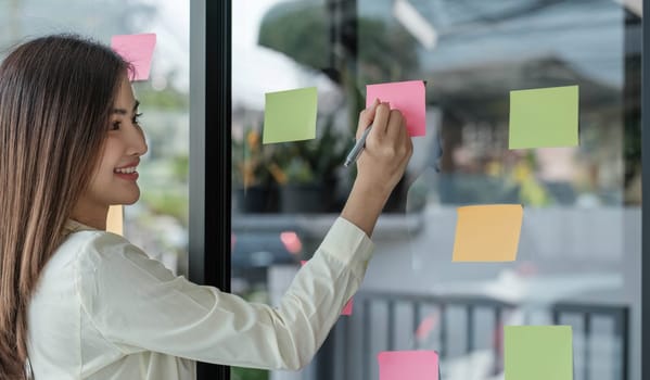 Businesswoman using sticky notes in glass wall while holding coffee cup to writing strategy...