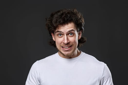 confused young bearded man in casual white t-shirt feels ashamed of terrible mistake on dark gray background. great idea