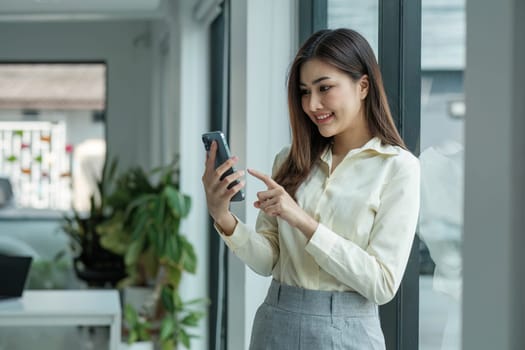 Smiling beautiful Asian businesswoman analyzing chart and graph showing changes on the market and holding smartphone at office