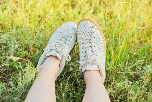 Close-up of female feet in white sneakers in green grass of summer meadow, top view