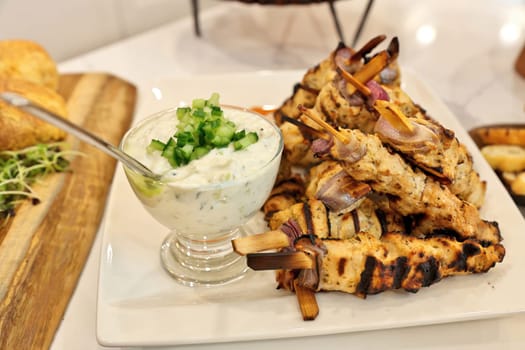 Grilled Greek chicken souvlaki skewers or kebabs piled high on a white plate with a bowl of tzatziki sauce.