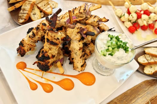 High angle view of Grilled Greek chicken souvlaki skewers or kebabs piled high on a white plate with a bowl of tzatziki sauce