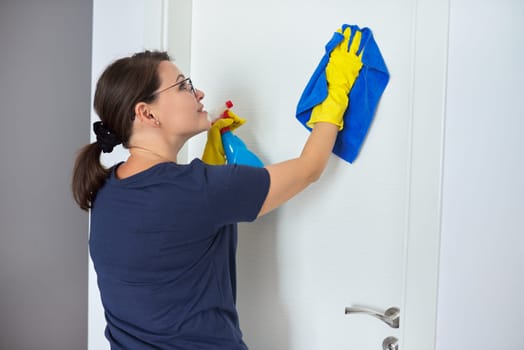 House cleaning, mature woman cleans at home in living room, in yellow gloves with spray, cleans white interior door. Hygiene, purity, cleanliness concept