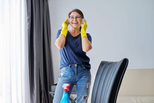 Mature positive woman in headphones rubber gloves doing house cleaning. People, housework and housekeeping concept