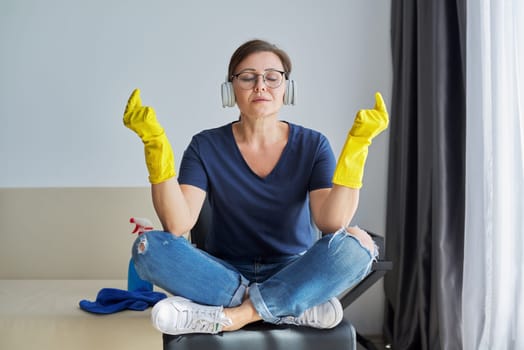 Sad tired meditating woman in headphones gloves with detergent and rag in room finished cleaning house. Hygiene, purity, home, cleanliness concept