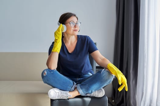 Mature positive woman in headphones rubber gloves doing house cleaning. People, housework and housekeeping concept