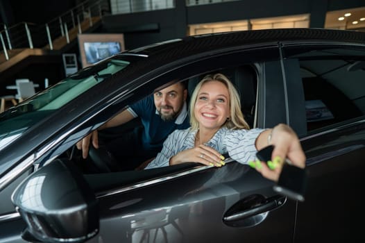 Happy caucasian couple is sitting in a car in a car dealership. A woman holds the keys to a new car