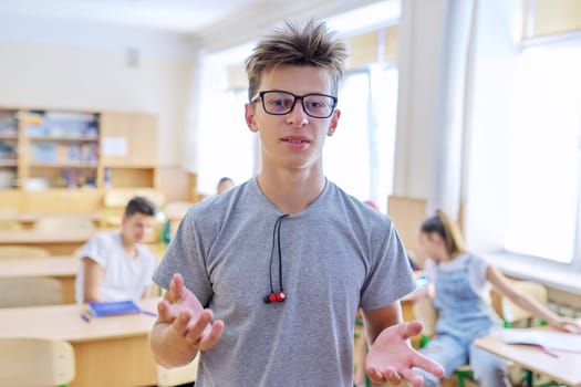 Talking gesturing student teenager at lesson in classroom, smiling handsome male with glasses looking at camera, background students sitting at desks