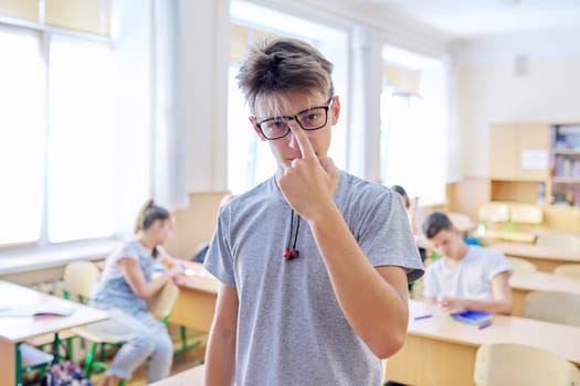 Smart teenager student male taking exam, answers lesson, talking looking at camera, classroom with study students background. High school, education, youth concept